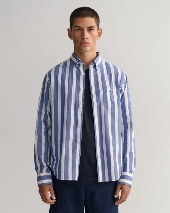 A young person with short hair stands facing forward, wearing a blue and white vertical striped button-up shirt over a dark t-shirt and dark pants, against a plain light background.