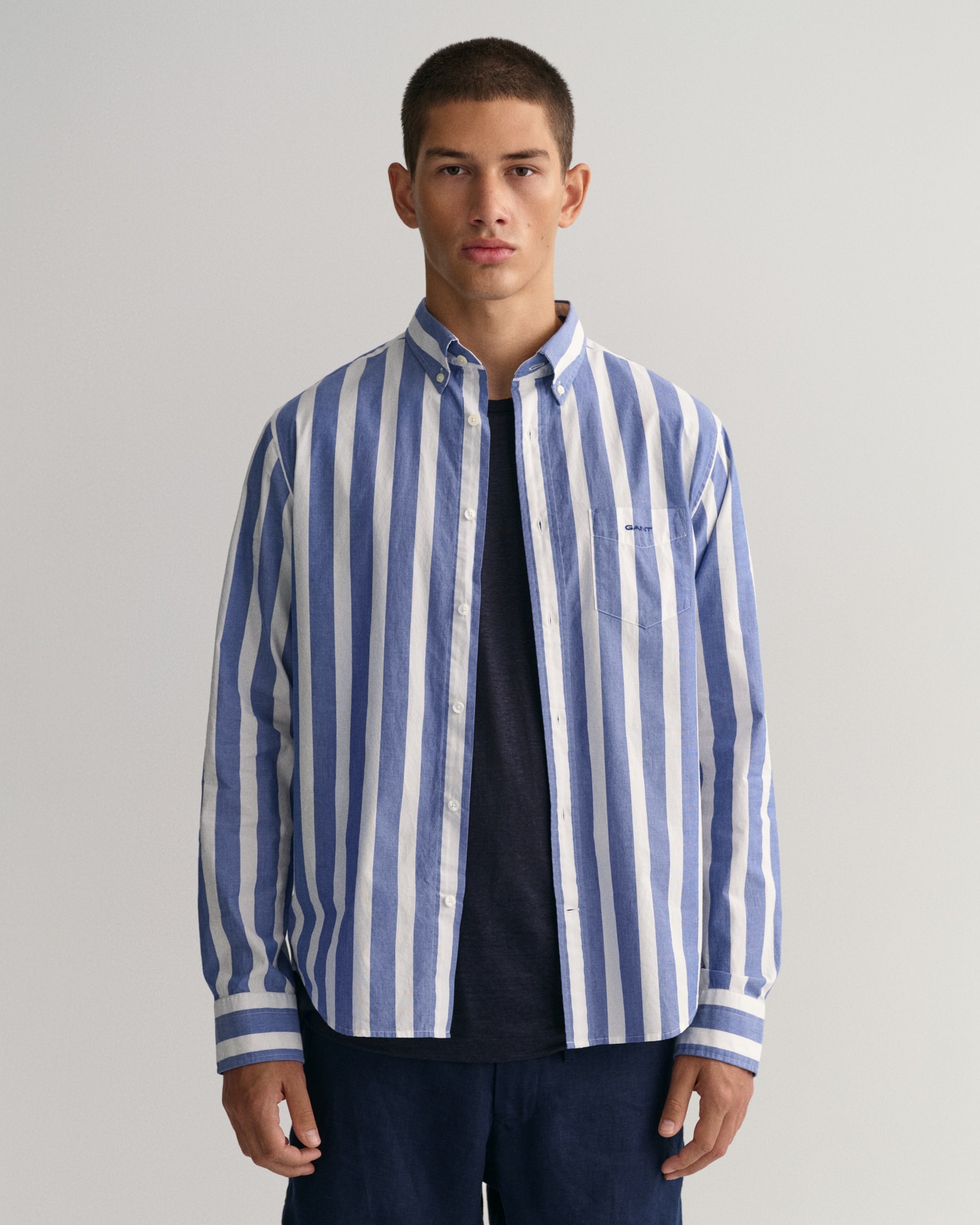 A young person with short hair stands facing forward, wearing a blue and white vertical striped button-up shirt over a dark t-shirt and dark pants, against a plain light background.