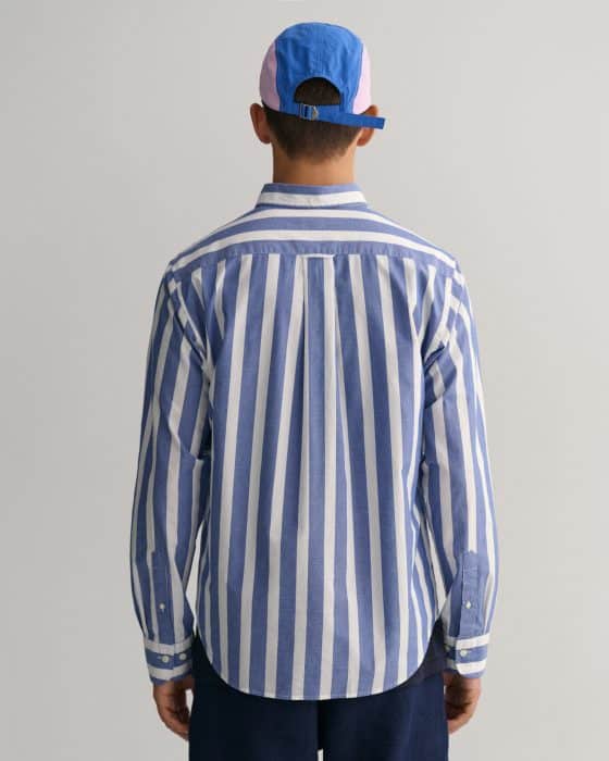 A person stands facing away, wearing a blue and white vertically striped button-up shirt, navy pants, and a blue and pink baseball cap against a plain light gray background.