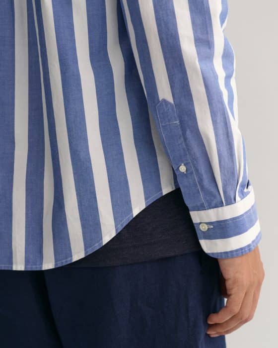A person wearing a blue and white vertical striped shirt with buttoned cuffs, dark blue pants, and a dark shirt underneath. The image focuses on the side and sleeve of the shirt and the persons hand.