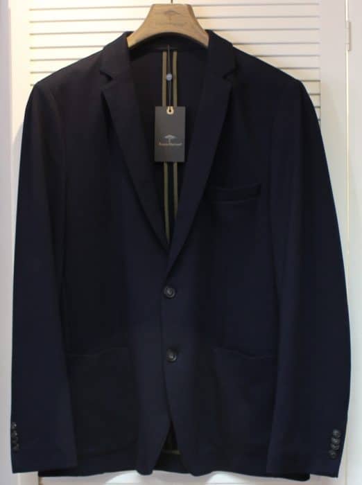 A dark navy blue suit jacket on a wooden hanger, displayed against a white louvered background, with a black tag hanging from the collar.
