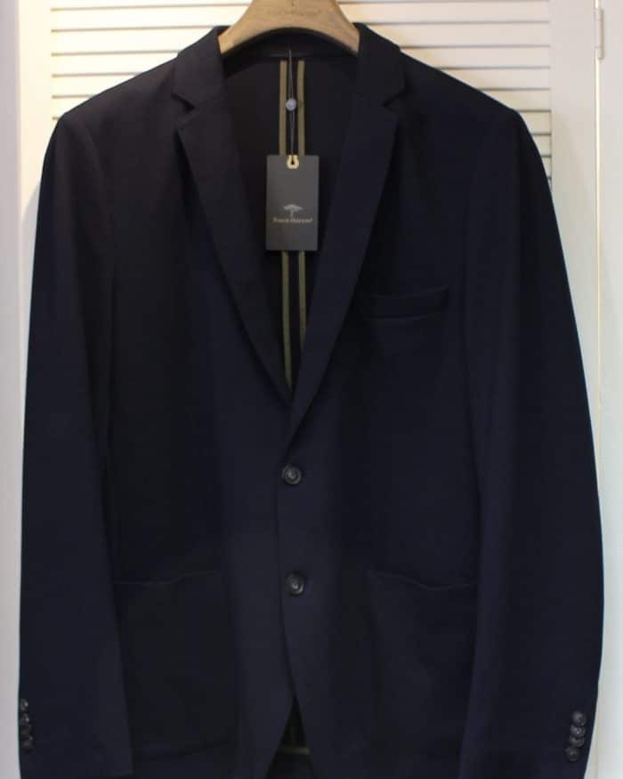 A dark navy blue suit jacket on a wooden hanger, displayed against a white louvered background, with a black tag hanging from the collar.