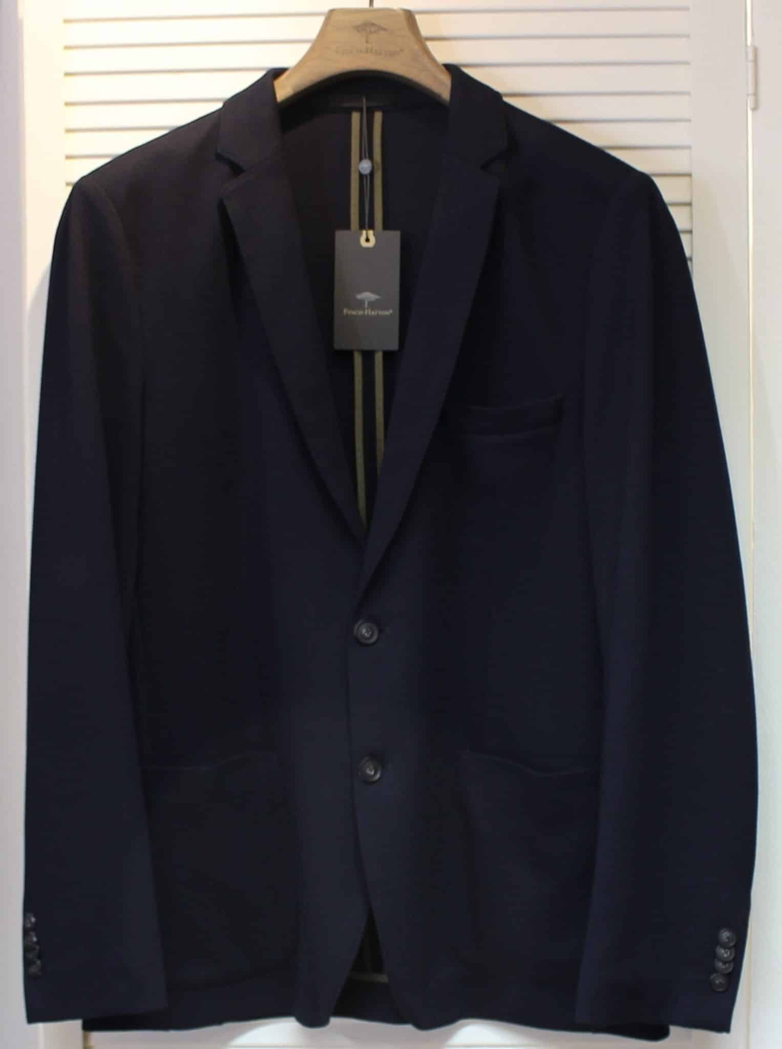 A dark navy blue suit jacket on a wooden hanger, displayed against a white louvered background, with a black tag hanging from the collar.