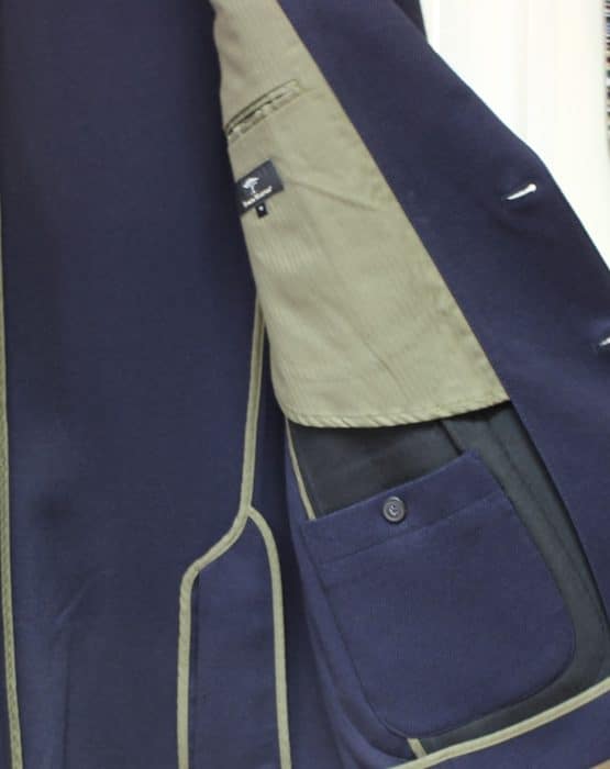 Close-up of the inside of a navy blue blazer with olive green lining and trim, showing an interior pocket with a button and a clothing label above the pocket.