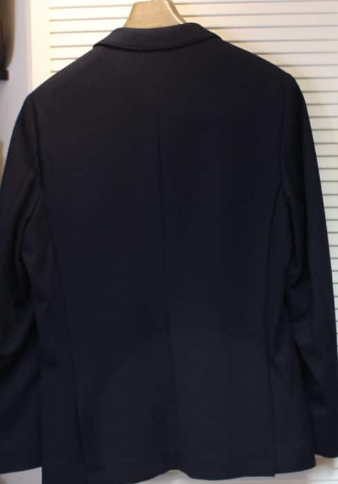 A navy blue blazer is displayed on a hanger, shown from the back against a white, slatted background.