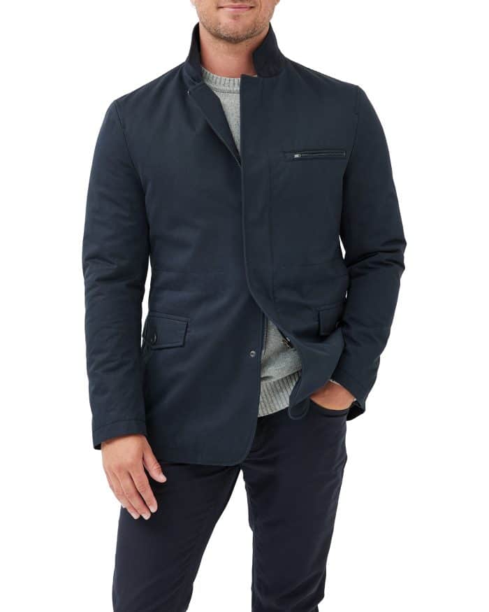 A man wearing a dark navy blazer over a light gray sweater and dark pants stands with one hand in his pocket against a white background.