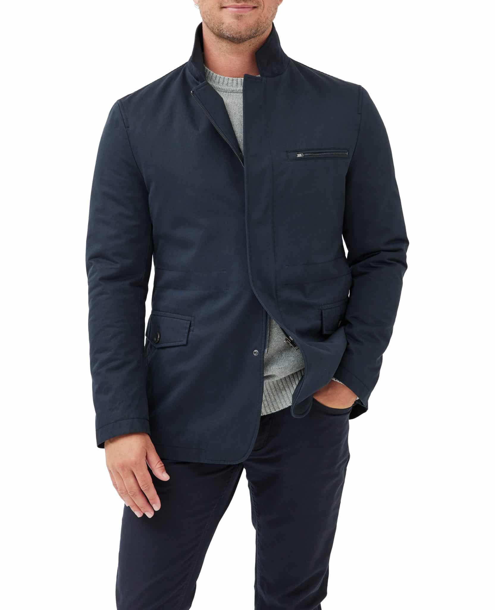 A man wearing a dark navy blazer over a light gray sweater and dark pants stands with one hand in his pocket against a white background.