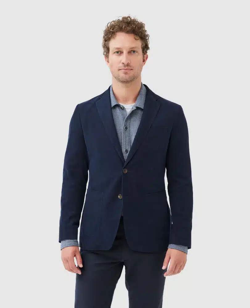 A man with short curly hair wearing a navy blue blazer over a blue checkered shirt and dark pants stands facing forward against a plain light gray background.