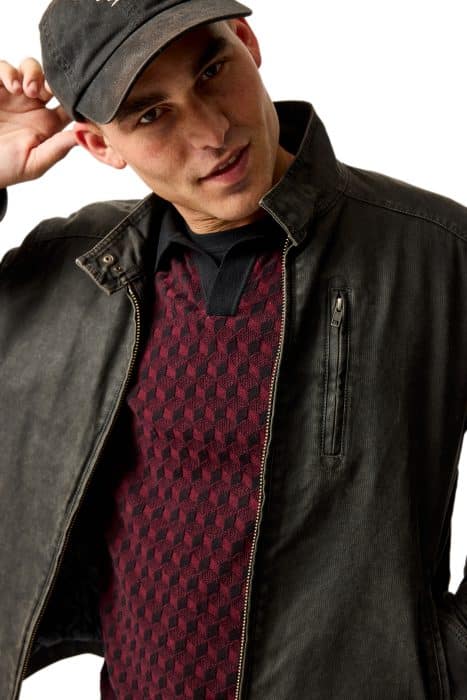 A man wearing a black baseball cap, black jacket, and a red patterned sweater looks at the camera while tilting his head and adjusting his cap, standing against a white background.