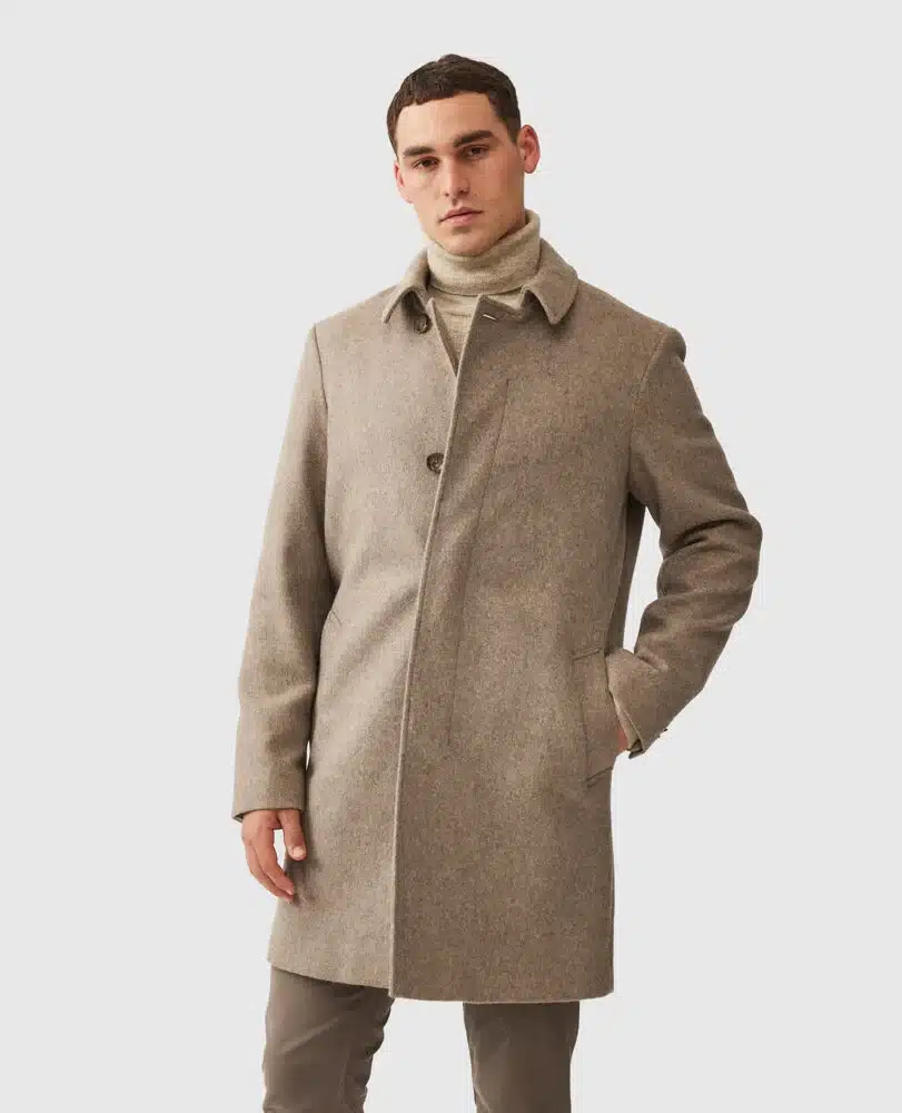 A man wearing a light brown overcoat, beige turtleneck sweater, and brown pants stands with one hand in his pocket against a plain light background.