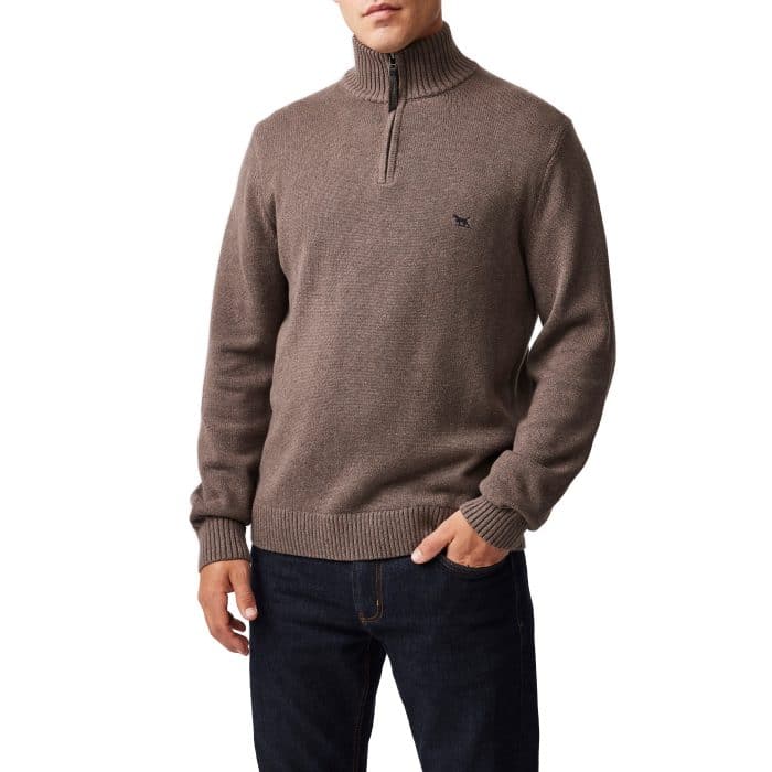 A man wearing a brown quarter-zip sweater with ribbed cuffs and collar, paired with dark blue jeans. His left hand is in his pocket and his right arm is relaxed by his side.