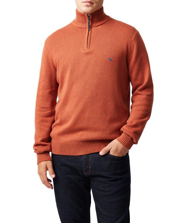 A person wearing an orange quarter-zip sweater with ribbed cuffs and hem, paired with dark blue jeans, standing against a plain white background.