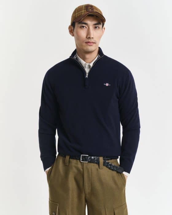 A man wearing a navy quarter-zip sweater over a beige shirt, olive green pants, a woven belt, and a plaid baseball cap stands against a plain white background.