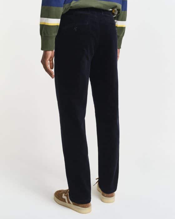 A person is standing and facing away from the camera, wearing dark navy corduroy pants, a green and blue striped sweater, and tan suede sneakers with white soles.