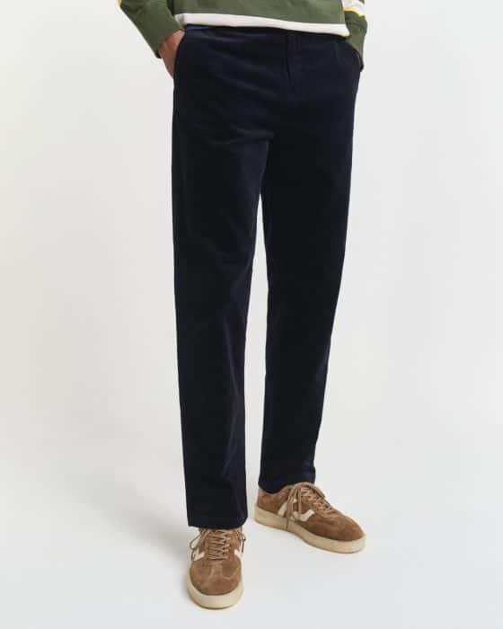 A person wearing dark navy corduroy pants, a green and white sweater, and brown suede sneakers with white soles, standing against a plain white background. Only the lower body is visible.