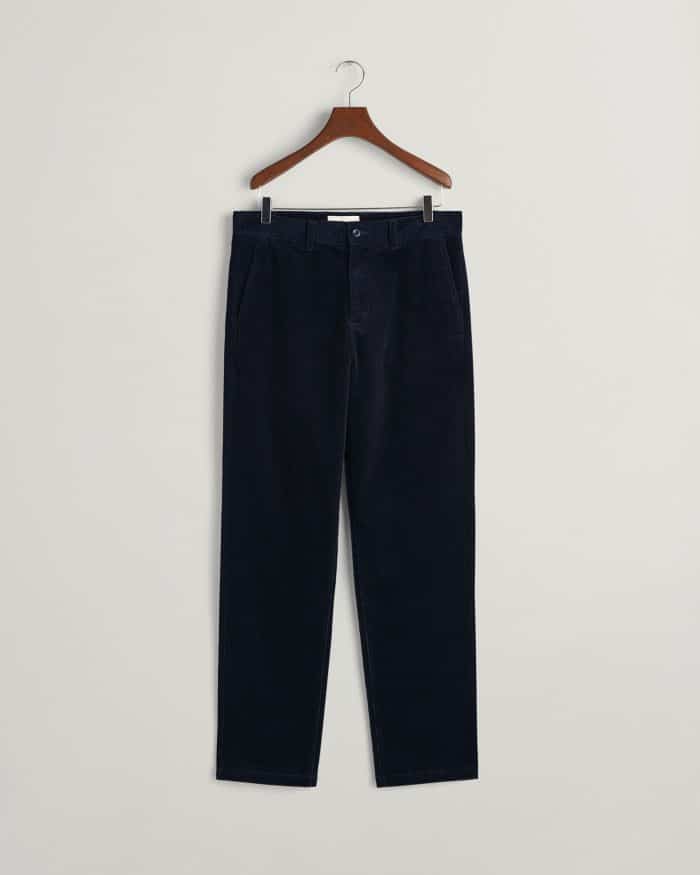 A pair of dark navy blue corduroy trousers hanging on a wooden hanger against a plain light gray background.
