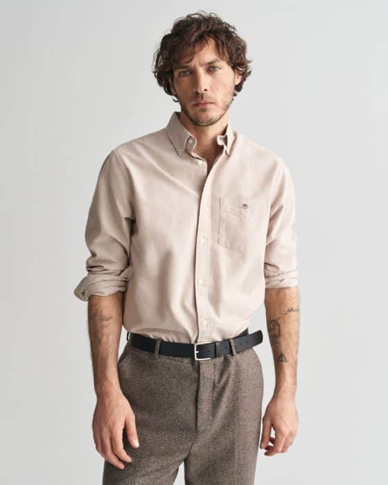 A man with wavy brown hair and light facial hair wears a beige button-up shirt with rolled sleeves, brown pants, and a black belt. He stands against a plain light background, looking directly at the camera.