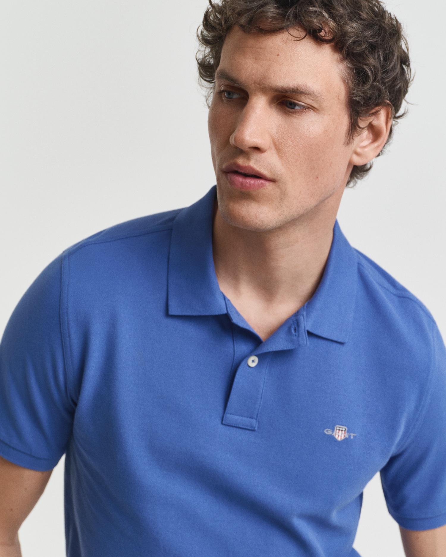A young man with short, curly brown hair wears a blue polo shirt with a small embroidered logo on the chest, looking slightly to his left against a plain white background.