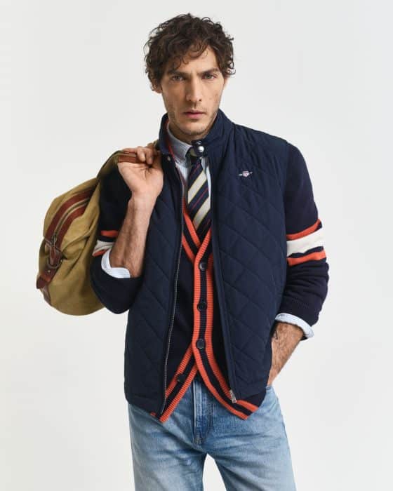 A man with curly hair wears a navy quilted vest over a striped cardigan and shirt with a tie, holding a tan duffel bag over his shoulder, posing against a plain white background.