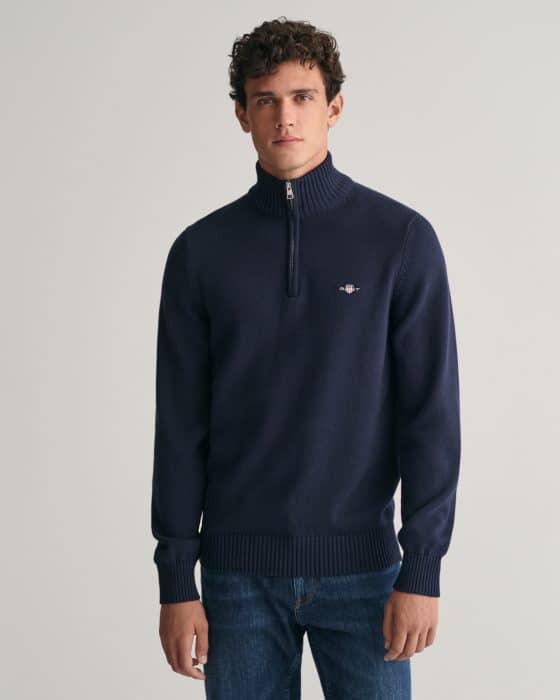 A young man with short curly hair wears a navy blue quarter-zip sweater with a small logo on the chest and blue jeans, standing against a plain light gray background.