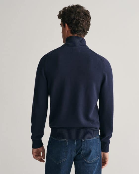 Man standing with his back to the camera, wearing a dark blue turtleneck sweater and blue jeans against a plain light background.
