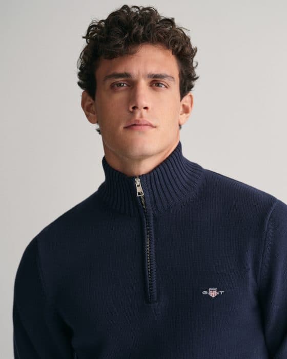 A man with short curly brown hair wearing a navy blue half-zip sweater with a stand-up collar and a small embroidered logo on the chest, posing against a neutral background.