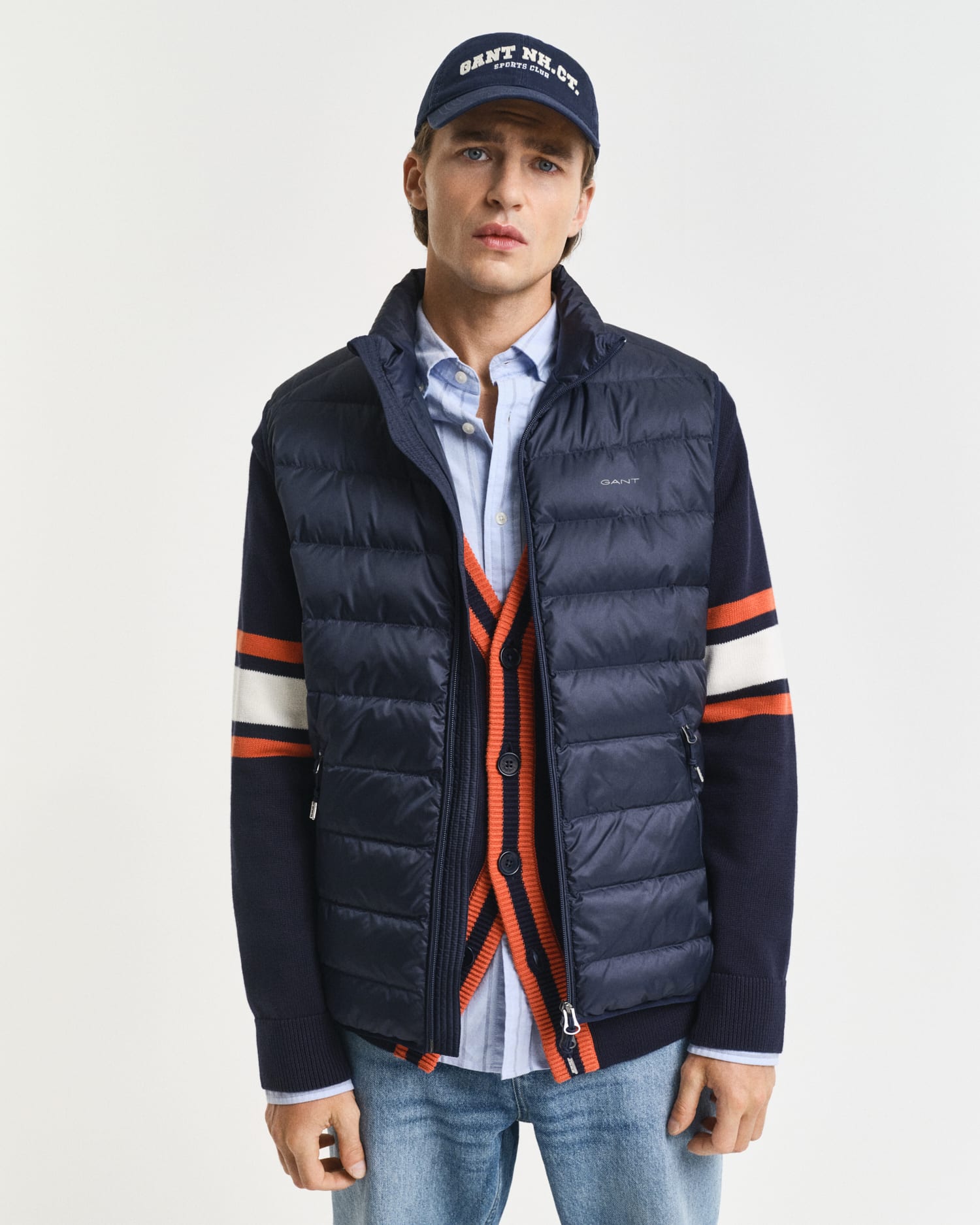 A young man wearing a navy puffer vest over a navy cardigan with orange and white stripes, a blue button-up shirt, jeans, and a navy cap stands against a plain white background.
