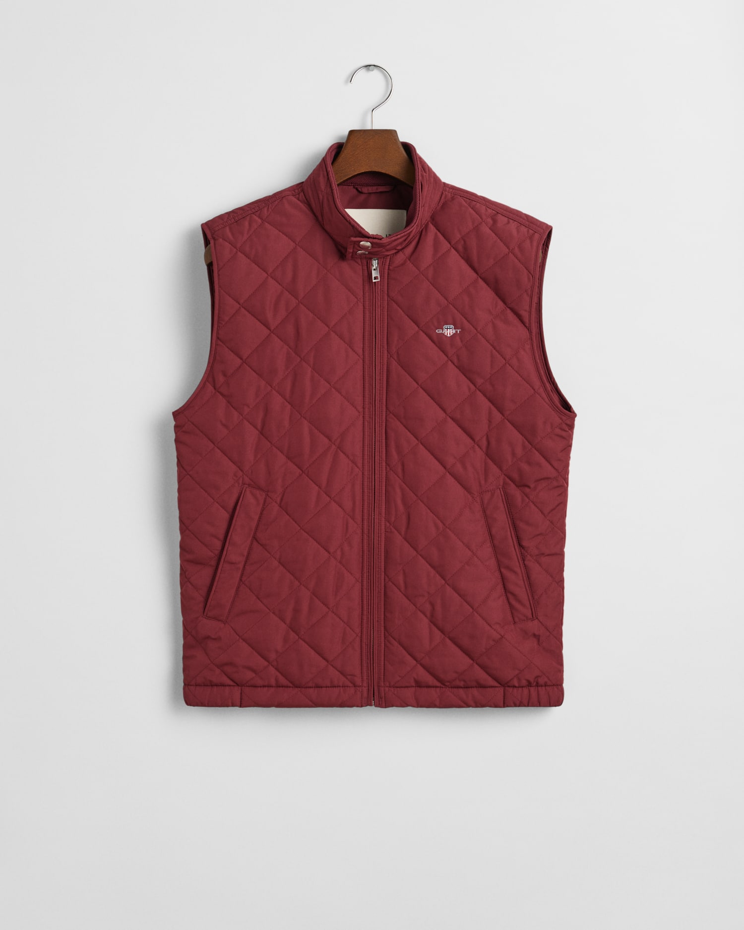 The GANT Quilted Windcheater Gilet is a sleeveless maroon gilet with a front zip, two slanted zipped pockets, and a small embroidered logo on the left chest, shown hanging on a wooden hanger against a white background.