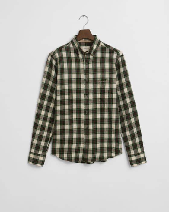 A long-sleeved, button-up flannel shirt with a green, black, and white plaid pattern hangs on a wooden hanger against a plain white background.