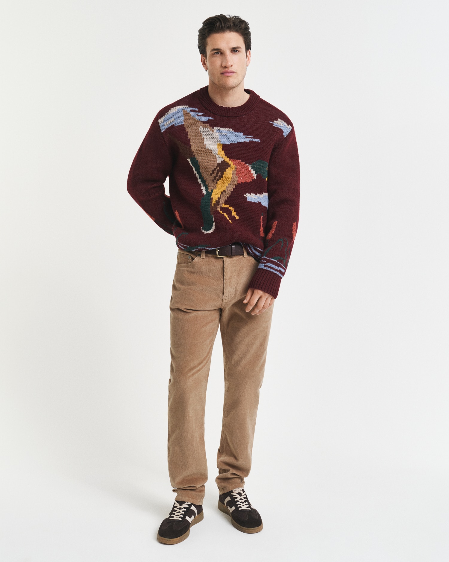 A man stands against a plain white background wearing a maroon sweater with an abstract dinosaur design, beige corduroy pants, and black sneakers. He has his left hand in his pocket and faces the camera.