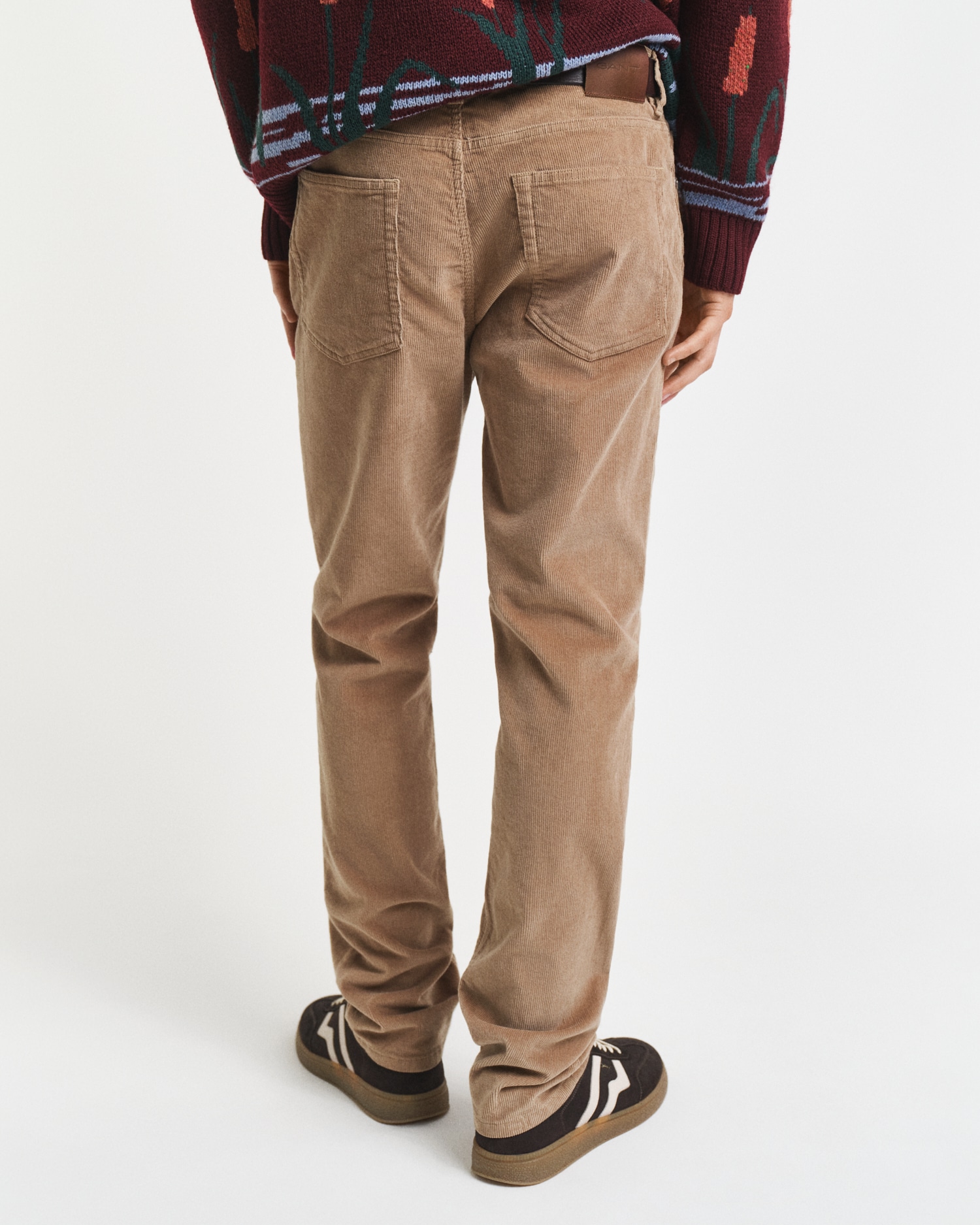 A person wearing light brown corduroy pants, a patterned sweater with maroon and green details, and black sneakers with white stripes, standing facing away from the camera against a plain white background.
