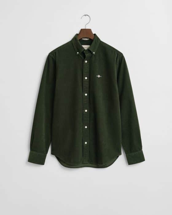 A long-sleeve, button-up, dark green corduroy shirt with a collar, white buttons, and a small embroidered logo on the left chest, hanging on a wooden hanger against a white background.