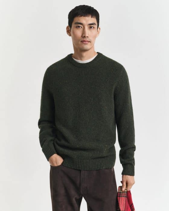 A man with short black hair wearing a dark green sweater and brown pants stands against a plain white background, holding a red plaid fabric in his left hand.