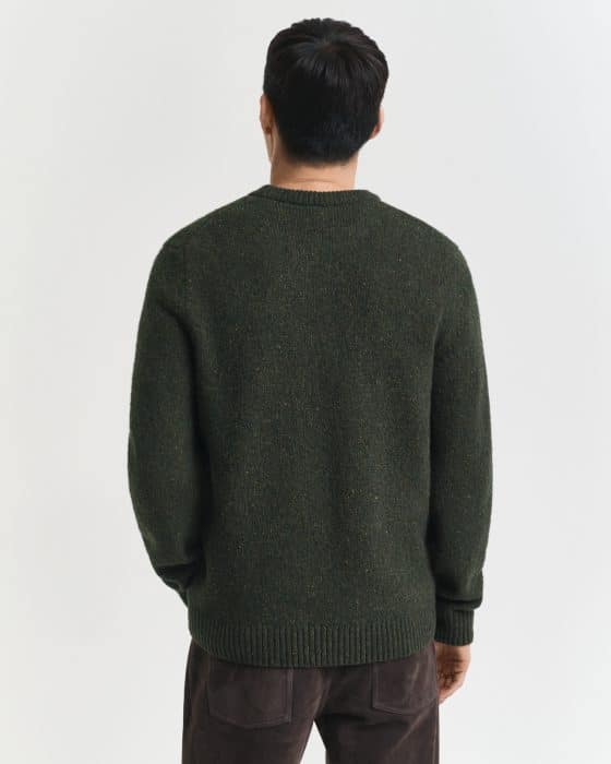 A person with short dark hair stands facing away, wearing a dark green knitted sweater and dark brown pants against a plain white background.