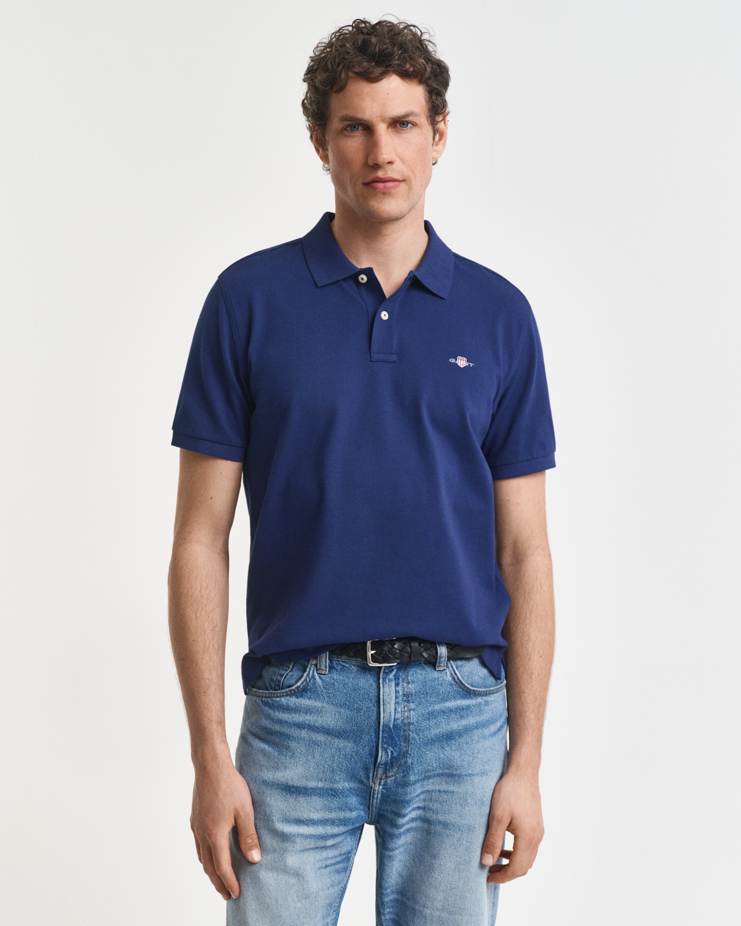 A man with short curly hair wearing a navy blue polo shirt and light blue jeans stands facing the camera against a plain white background.