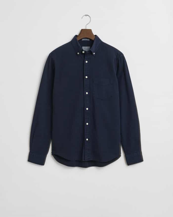 A navy blue long-sleeve button-up shirt with a front pocket, button-down collar, and white buttons, hanging on a wooden hanger against a white background.