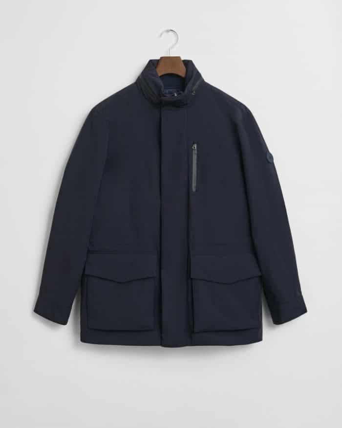 A navy blue, long-sleeve jacket with a high collar, zippered chest pocket, two large front flap pockets, and a subtle logo patch on the upper left sleeve, displayed on a hanger against a white background.