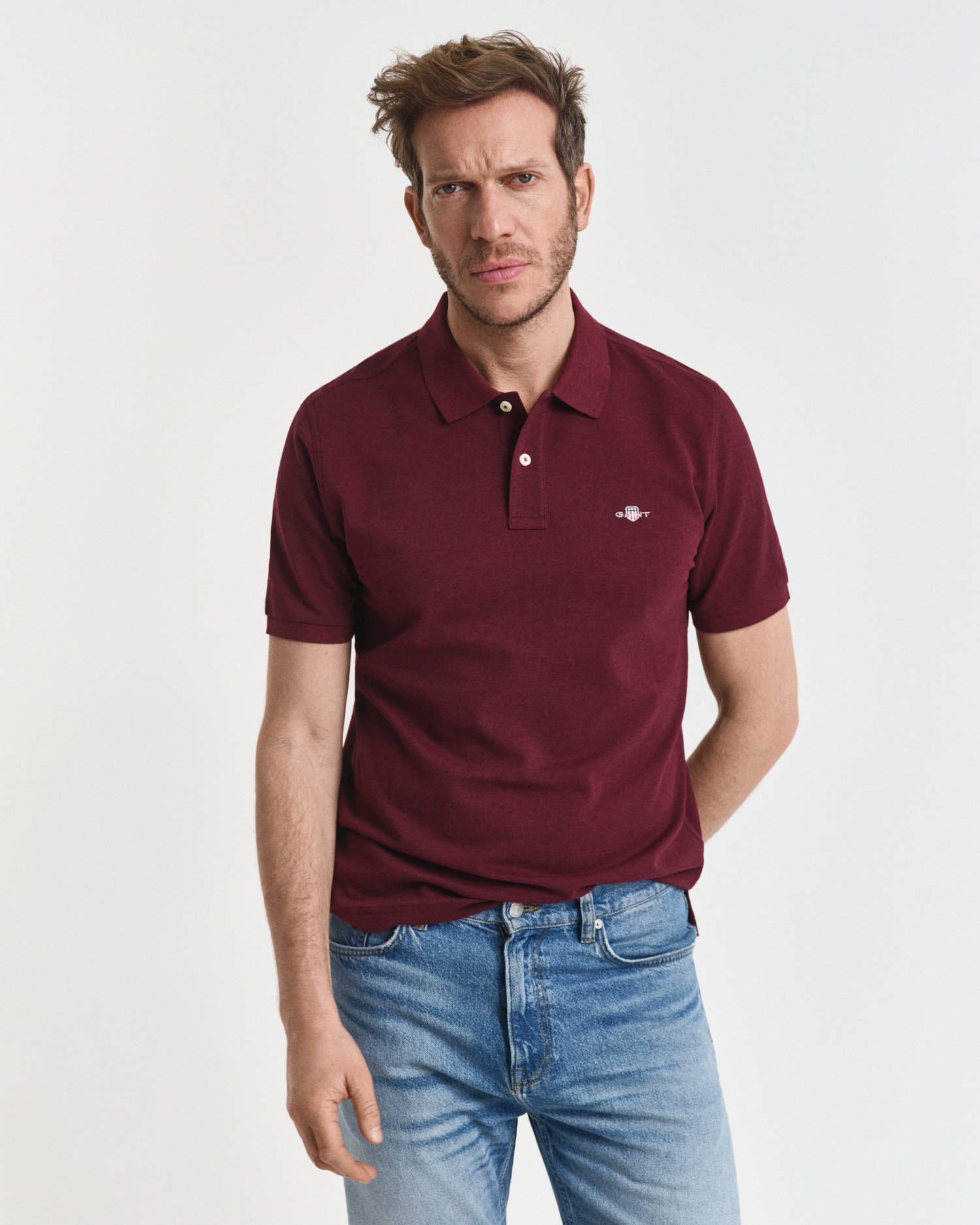 A man with short brown hair and light stubble wears a burgundy polo shirt and light blue jeans, standing against a plain white background. He has a neutral expression and his left hand is in his pocket.