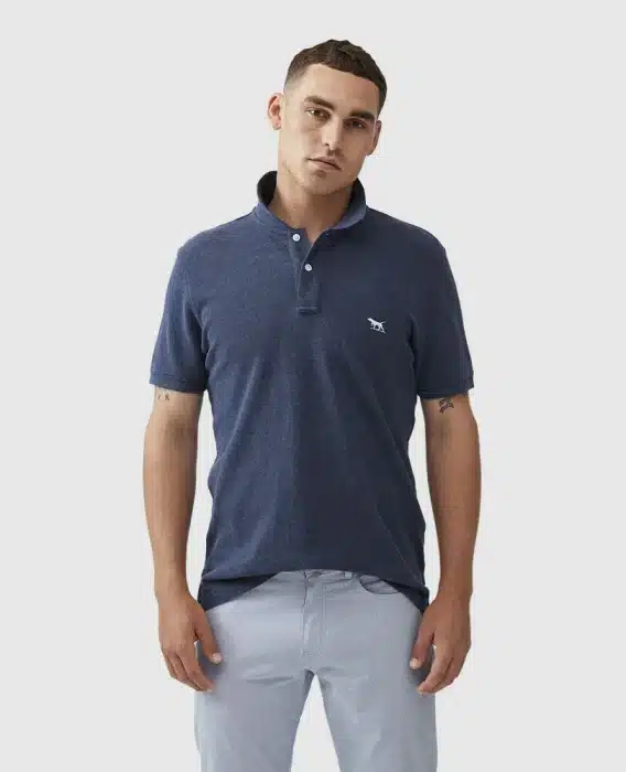 A man with short brown hair wears a navy blue polo shirt with a small embroidered logo and light gray pants, standing against a plain light background with a relaxed expression.