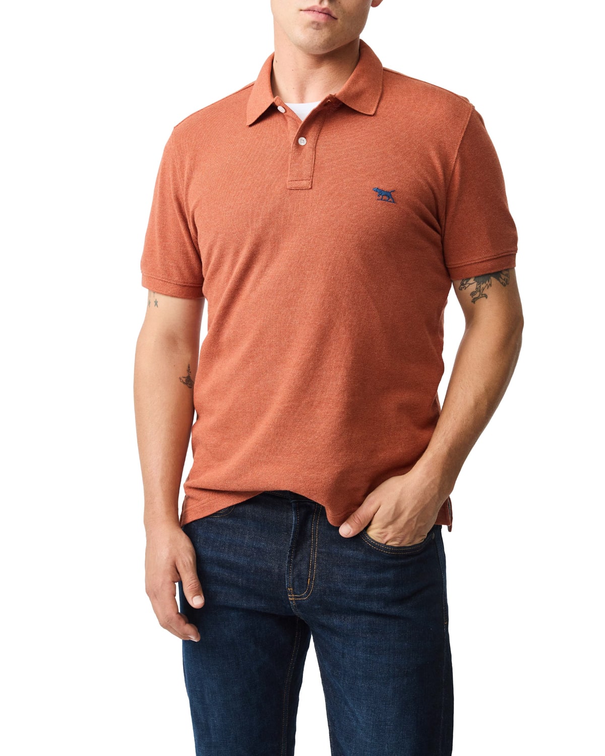 A man wearing a rust-colored short-sleeve polo shirt and dark jeans stands with one hand in his pocket against a plain white background.