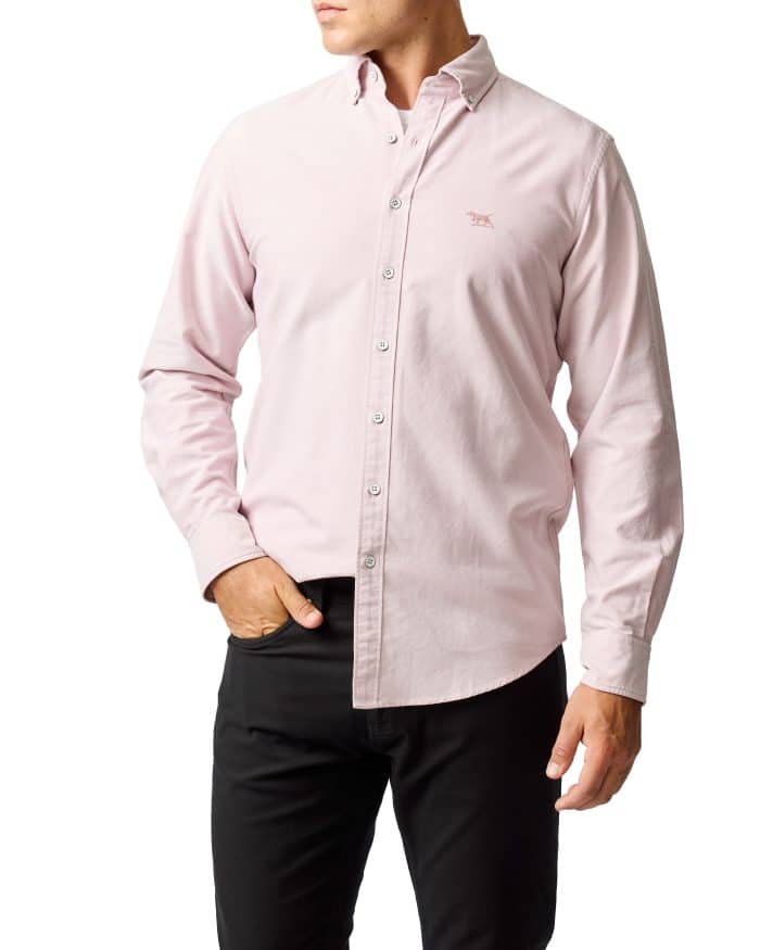 A man wearing a light pink button-down shirt with long sleeves and black pants, standing with one hand in his pocket against a white background.