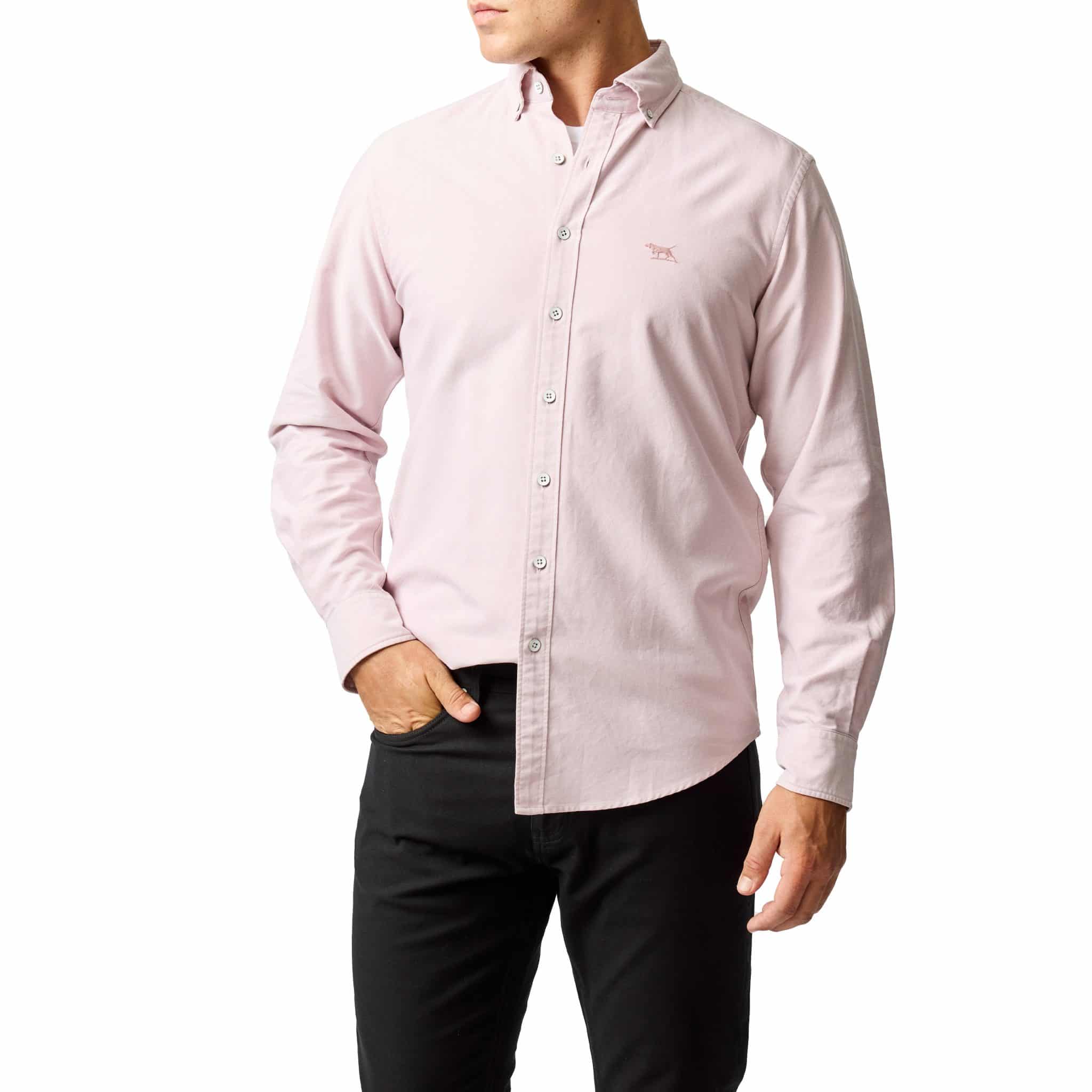 A man wearing a light pink button-down shirt with long sleeves and black pants, standing with one hand in his pocket against a white background.