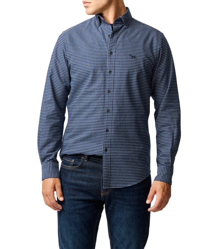 A person wearing a blue and black checkered long-sleeve button-down shirt and dark blue jeans, standing against a white background. The persons head is turned slightly to the side.