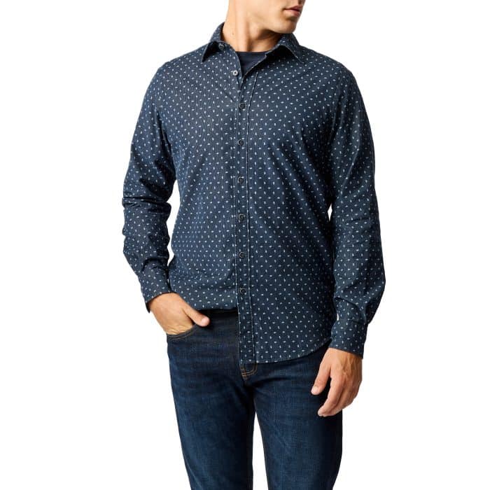 A man wearing a navy blue button-up shirt with a small white geometric pattern and dark blue jeans, standing with one hand in his pocket against a white background.
