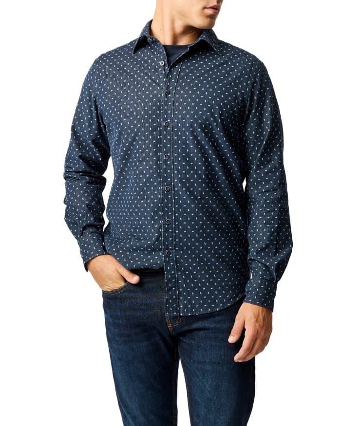A man wearing a navy blue button-up shirt with a small white geometric pattern and dark blue jeans, standing with one hand in his pocket against a white background.