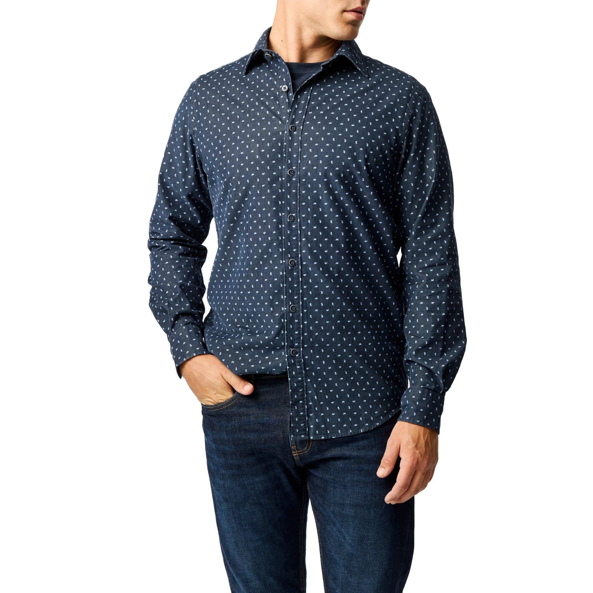 A man wearing a navy blue button-up shirt with a small white geometric pattern and dark blue jeans, standing with one hand in his pocket against a white background.