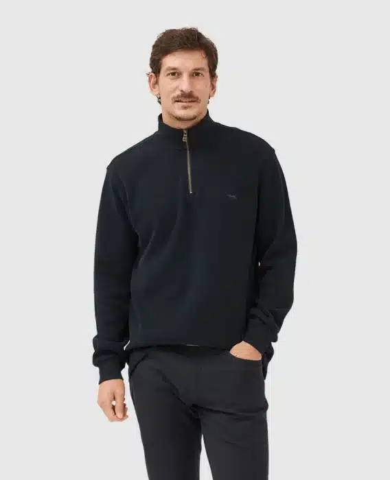 A man with short brown hair and a mustache wears a black quarter-zip pullover and black pants, standing against a plain light gray background with one hand in his pocket.