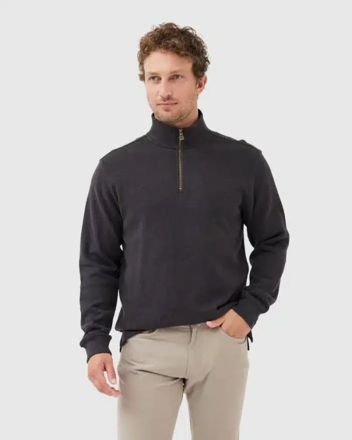 A man with curly hair wearing a dark quarter-zip sweater and light beige pants stands against a plain light gray background, with one hand in his pocket.