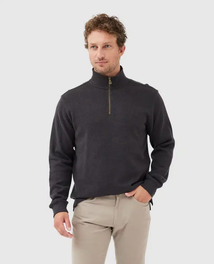 A man with curly hair wearing a dark quarter-zip sweater and light beige pants stands against a plain light gray background, with one hand in his pocket.