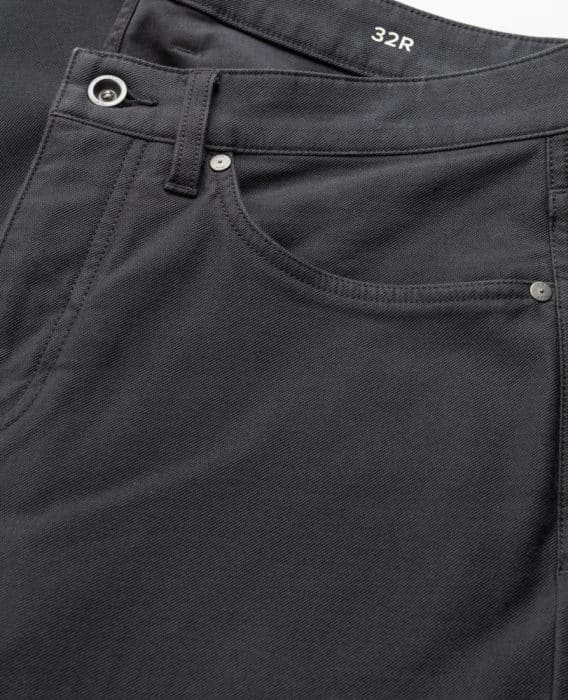 Close-up of the front pocket area of a pair of black jeans with visible stitching, metal rivets, a belt loop, and a button. The waistband is marked with the size 32R in white text.